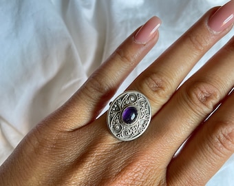 May include: A silver ring with an ornate design and a purple gemstone in the center.