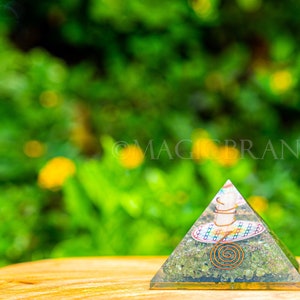 May include: A green, brown, and white crystal pyramid with a copper wire spiral design. The pyramid is sitting on a wooden surface with a blurred green background.