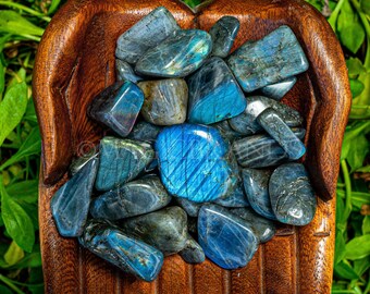 May include: A collection of tumbled labradorite stones in various shades of blue and grey, displayed in a wooden hand-shaped bowl. The stones have a shimmering iridescent effect.