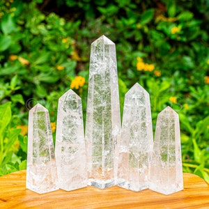 May include: Six clear quartz crystal points arranged in a row on a wooden surface. The points are all different sizes and shapes, but they are all clear and have a natural, unpolished look.