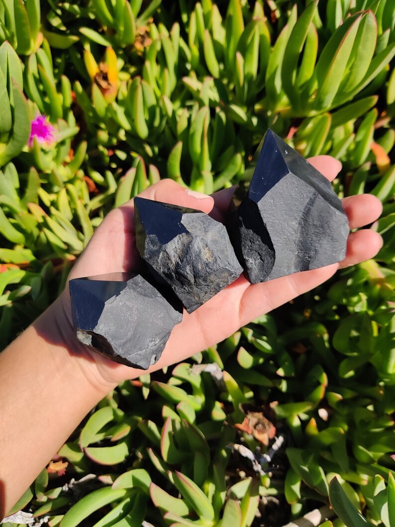May include: Three black obsidian crystal points, also known as black quartz, are held in a hand. The crystals are rough and have a natural, unpolished look.
