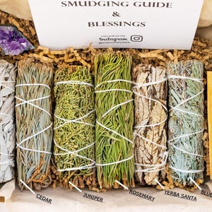 May include: A set of six smudging sticks in a box with a white card that says "Smudging Guide & Blessings". The sticks are white sage, cedar, juniper, rosemary, yerba santa, and blue sage. There is also a small amethyst crystal and a palo santo stick.