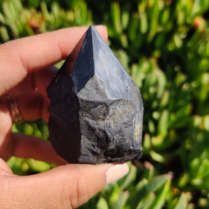 May include: A black obsidian crystal point with a rough, natural texture. The crystal is pointed at the top and has a flat base.