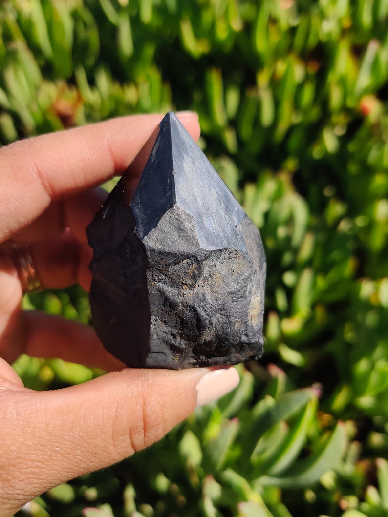 May include: A black obsidian crystal point with a rough, natural texture. The crystal is pointed at the top and has a flat base.