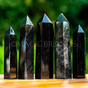 May include: Six black obsidian crystal points of varying sizes, arranged in a row on a wooden surface. The crystals are smooth and polished, with a dark, almost black color.