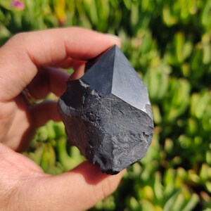 May include: A black obsidian crystal point with a rough, natural texture. The crystal is shaped like a pyramid with a pointed tip.