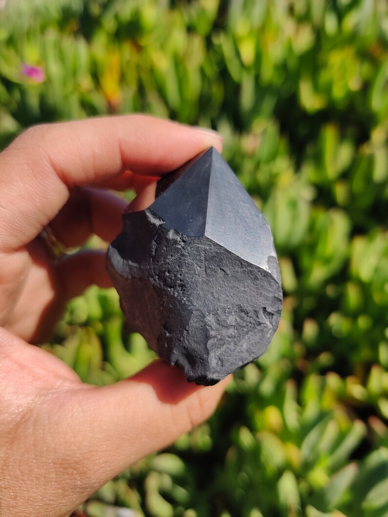May include: A black obsidian crystal point with a rough, natural texture. The crystal is shaped like a pyramid with a pointed tip.