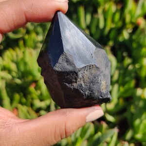 May include: A black obsidian crystal point with a faceted, rough-cut design. The crystal is held in a hand against a green background.
