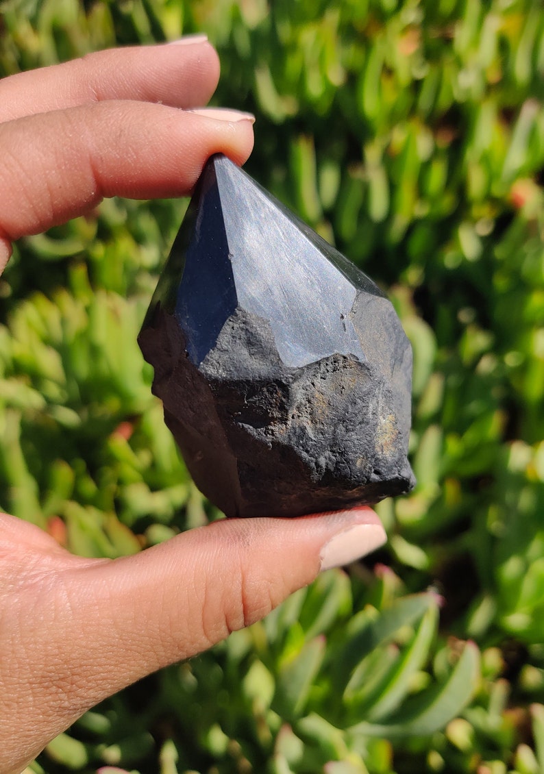 May include: A black obsidian crystal point with a faceted, rough-cut design. The crystal is held in a hand against a green background.