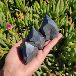 May include: Three black obsidian crystal points, also known as black quartz, are held in a hand. The crystals are sharp and have a dark, glassy appearance.