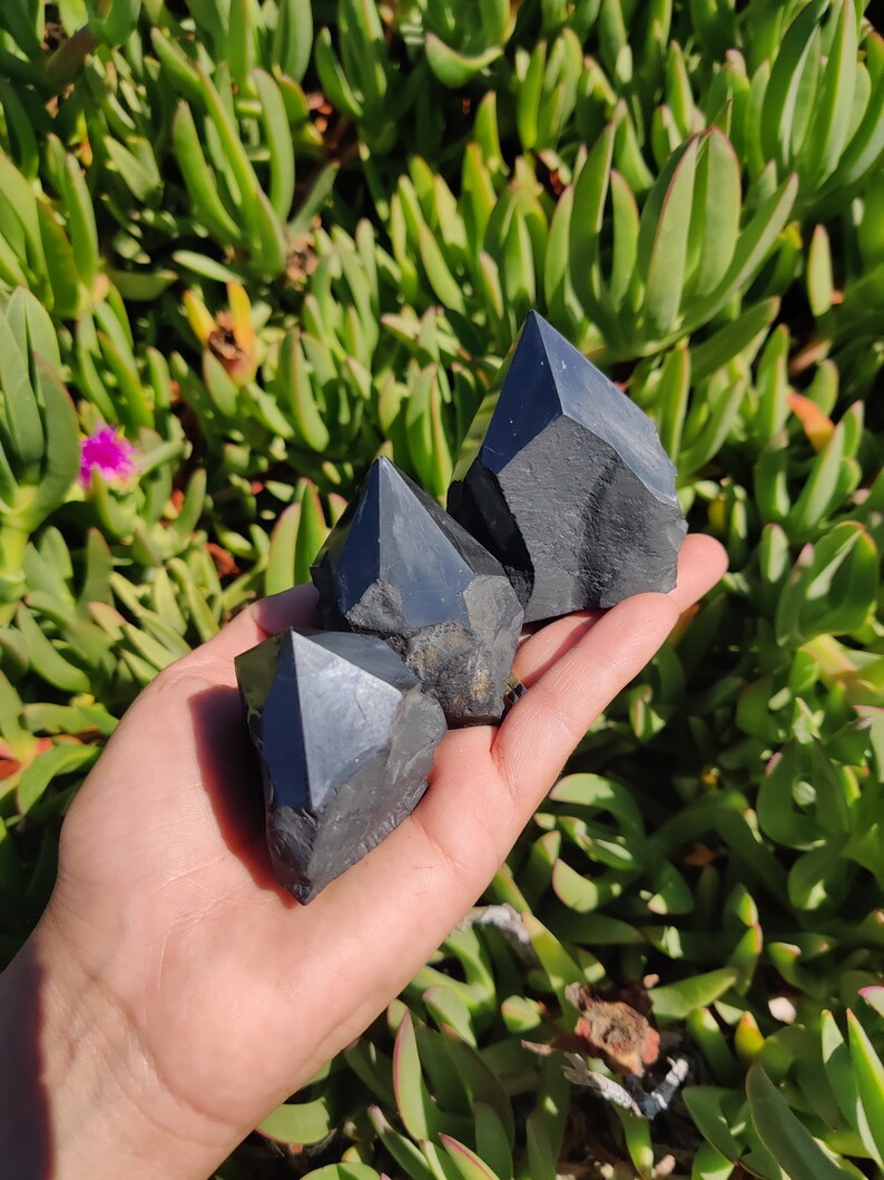 May include: Three black obsidian crystal points, also known as black quartz, are held in a hand. The crystals are sharp and have a dark, glassy appearance.
