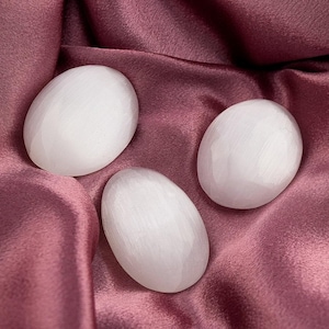 May include: Three smooth, oval-shaped white stones rest on a draped, deep rose-colored fabric. The stones have a polished appearance, and the fabric has a soft, satiny texture. The composition creates a serene and elegant aesthetic.
