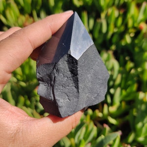 May include: Black obsidian crystal point, a dark, smooth, and pointed stone with a rough, textured base. The crystal is held in a hand against a green background.