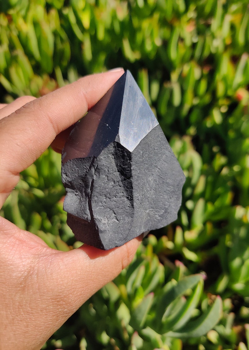 May include: Black obsidian crystal point, a dark, smooth, and pointed stone with a rough, textured base. The crystal is held in a hand against a green background.