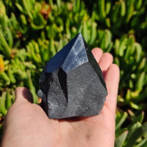 May include: A black obsidian crystal point with a rough, natural texture. The crystal is shaped like a pyramid and has a sharp point. It is held in a hand against a green background.