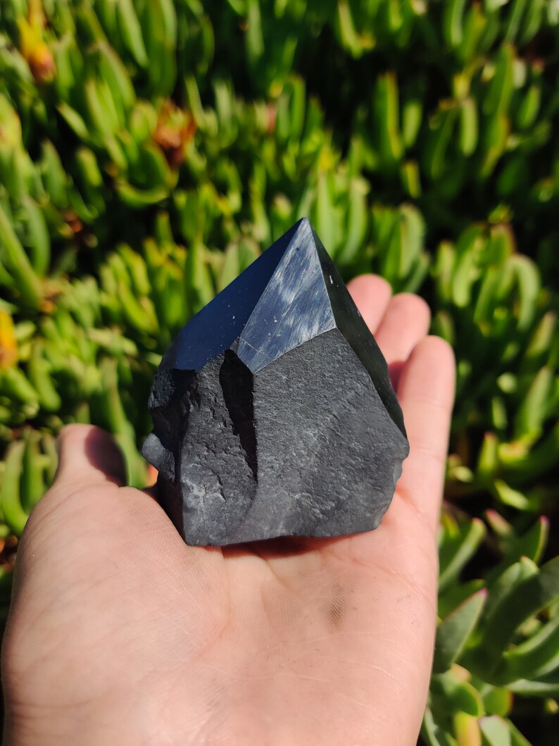 May include: A black obsidian crystal point with a rough, natural texture. The crystal is shaped like a pyramid and has a sharp point. It is held in a hand against a green background.