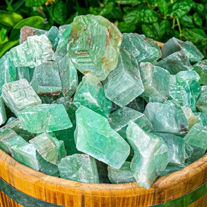 May include: A wooden barrel filled with numerous rough-cut, translucent green crystals. The crystals have a crystalline structure and varying shades of green. The barrel is made of light-colored wood and is surrounded by green foliage.