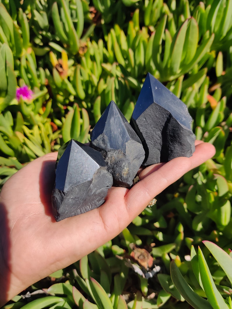 May include: Three black obsidian crystal points in various sizes, arranged in a triangular formation, held in a hand against a green leafy background.