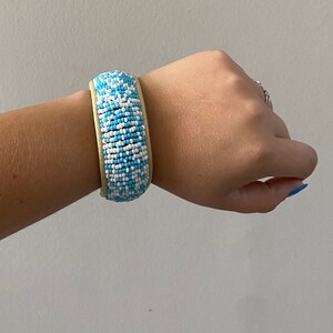 Puede incluir: Un brazalete de cuentas azules y blancas con un respaldo de cuero marrón claro.