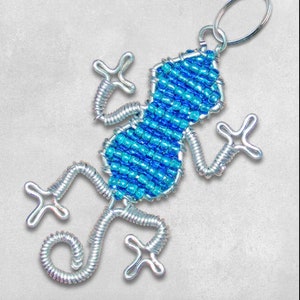 Op de afbeelding: Een blauwe en zilveren kralen gecko sleutelhanger. De gecko is gemaakt van draad en heeft een zilveren sleutelring.