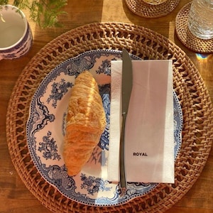 May include: A blue and white patterned plate with a croissant on top. The plate is on a woven rattan placemat with a white napkin and a silver knife. The text "ROYAL" is printed on the napkin.
