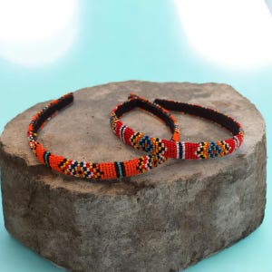 May include: Two thin headbands featuring intricate beadwork in vibrant orange, red, blue, yellow, and black. The beads are arranged in geometric patterns along the entire length of the bands, which have a black interior lining for a secure fit.