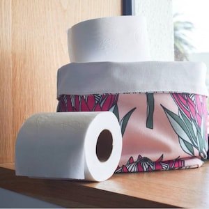 Puede incluir: Una cesta de almacenamiento de tela con estampado floral rosa y verde con un forro de tela blanco. Dos rollos de papel higiénico blanco están encima de la cesta.