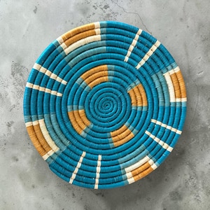 May include: A round, flat, coiled basket featuring a vibrant pattern of concentric circles. The design incorporates alternating bands of bright turquoise, deep teal, warm mustard yellow, and cream white fibres woven into a tight, circular structure.