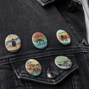May include: A black denim jacket with five oval-shaped pins. The pins feature colorful illustrations of animals and landscapes. The pins are attached to the jacket's front pocket and lapel, adding a decorative touch.
