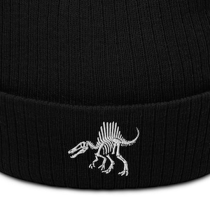 May include: Black ribbed knit beanie featuring a white Spinosaurus skeleton design. The beanie has a folded cuff and a classic, comfortable fit. This hat is a great accessory for anyone interested in paleontology or dinosaurs.