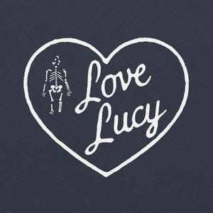 May include: White heart-shaped graphic with the text "Love Lucy" in cursive, and a small skeleton illustration on a dark blue background. The design is simple and clean, suitable for various products.