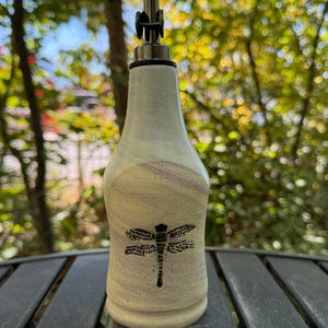 May include: A ceramic oil dispenser bottle with a cream-colored speckled glaze and a subtle marbled pattern. The bottle features a black dragonfly graphic on the front and is topped with a silver-colored metal pour spout with a black rubber seal.