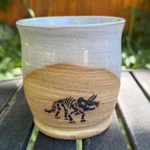 May include: A ceramic mug featuring a two-tone design with a smooth, light gray glazed upper section and a textured, natural tan clay base. A black, detailed illustration of a triceratops dinosaur skeleton is centered on the tan portion.