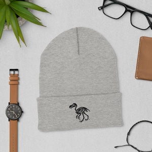 Könnte beinhalten: Hellgraue Mütze mit einem schwarzen, gestickten Dinosaurierskelett-Design. Die Mütze hat einen umgeschlagenen Saum und ist aus weichem Strickmaterial gefertigt. Die Mütze wird auf einer hellgrauen Oberfläche mit einer Uhr, einer Brille und einer Brieftasche gezeigt.