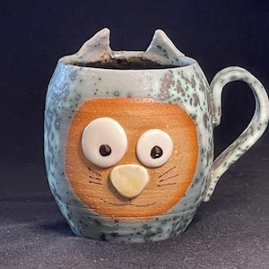 May include: A ceramic mug with a cat face design. The mug is light blue with brown spots and a brown cat face with white eyes and a yellow nose.