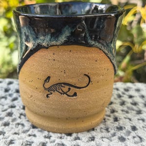 May include: A ceramic mug with a tan, speckled base and a dark blue, glossy glazed rim. The front of the mug features a small, black, stamped illustration of a dinosaur skeleton. The mug has a rounded body and a slightly flared, wide opening.