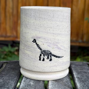 May include: A cylindrical ceramic tumbler with a speckled off-white glaze and subtle lavender marbling. The exterior features a black graphic illustration of a dinosaur skeleton, specifically a long-necked sauropod, centered on the front surface.