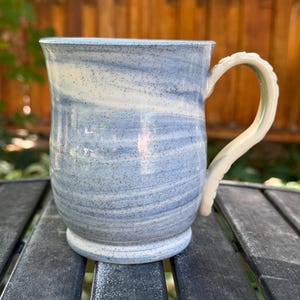 May include: A ceramic mug featuring a marbled pattern of light blue and white with dark speckles throughout. The mug has a wide, rounded body, a flared rim, and a textured, curved handle with a series of small, raised bumps along its outer edge.