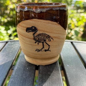 May include: A ceramic mug featuring a tan, unglazed lower section with a black line drawing of a dodo bird skeleton. The upper portion of the mug is finished with a glossy, dark brown glaze that drips slightly over the tan clay surface.