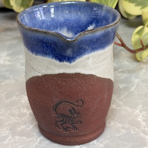 May include: A handmade ceramic pitcher with a three-tone design. The top is blue, the middle is white, and the bottom is a reddish-brown. A detailed illustration of a creature is etched into the reddish-brown section. The pitcher has a spout for easy pouring.