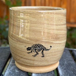 May include: A cylindrical ceramic mug with a smooth, glossy beige and tan marbled glaze. The exterior features a small, detailed black graphic of an ankylosaurus dinosaur skeleton positioned near the base of the vessel.