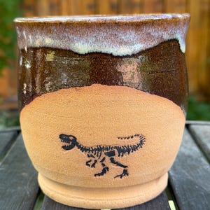 May include: A ceramic mug featuring a natural tan clay base with a black dinosaur skeleton illustration. The upper portion of the mug is glazed in a glossy dark brown with a lighter, creamy white drip effect along the rim and transition area.