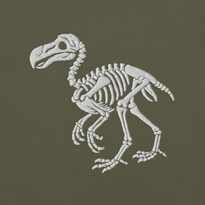 Könnte beinhalten: Weiß gesticktes Design eines Dodo-Vogelskeletts auf olivgrünem Hintergrund. Die detaillierte Stickerei zeigt den Schädel, den Brustkorb und die Knochen der Beine des Vogels. Der Dodo wird in einer Laufpose gezeigt.