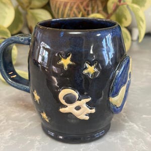 May include: A dark blue ceramic mug with a handle, decorated with yellow stars and a cream-colored astronaut figure. The mug also features a blue and yellow crescent moon design. The mug is a unique, handmade piece.