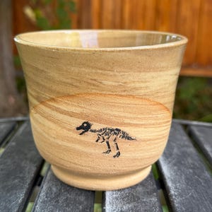 May include: A tan ceramic mug with a smooth, glossy finish and a wide, open top. The exterior features a small, detailed black illustration of a dinosaur skeleton positioned centrally on the side of the vessel, which has a slightly tapered base.