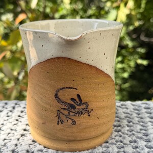 May include: A ceramic creamer pitcher featuring a two-tone design with a speckled cream-colored upper section and a raw, unglazed tan lower section. A black graphic illustration of a dinosaur skeleton is centered on the front of the pitcher.