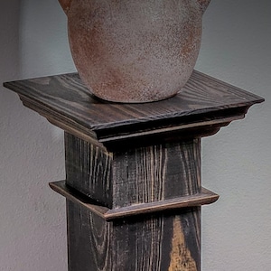 Puede incluir: Un pedestal rústico de madera teñida oscura con una parte superior e inferior cuadradas, que sostiene un jarrón de cerámica texturizado de color marrón claro. El diseño del pedestal presenta elementos en capas y una apariencia desgastada, adecuado para exhibir objetos decorativos.