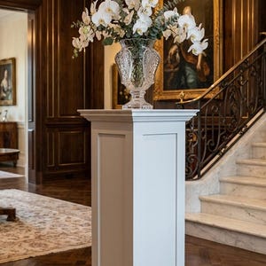 May include: A tall white rectangular pedestal with a flat top and base, supporting a clear, ornate cut-glass vase filled with white orchid flowers and green eucalyptus foliage, creating a structured display piece for interior spaces.