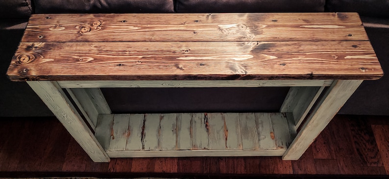 May include: A rustic wooden console table with a distressed white finish. The top is made of two planks of wood with a natural wood stain. The bottom shelf is also made of wood and has a distressed white finish.