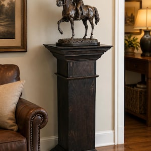 May include: A dark brown wooden pedestal with a square top and base, featuring a tiered molding design. Resting on the pedestal is a bronze-colored sculpture of a person in a military uniform riding a horse, set on a textured oval base.